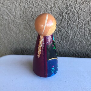 Peut inclure: Poup&eacute;e en bois peinte &agrave; la main repr&eacute;sentant un personnage avec une t&ecirc;te brun clair, une tresse blonde et une robe dans les tons de violet, noir et bleu. La poup&eacute;e a un design simple et stylis&eacute; avec des d&eacute;tails peints et est debout.