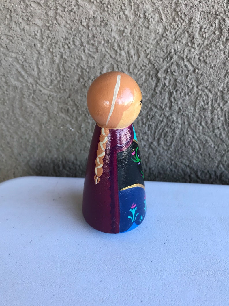 Peut inclure: Poup&eacute;e en bois peinte &agrave; la main repr&eacute;sentant un personnage avec une t&ecirc;te brun clair, une tresse blonde et une robe dans les tons de violet, noir et bleu. La poup&eacute;e a un design simple et stylis&eacute; avec des d&eacute;tails peints et est debout.