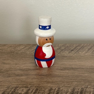 May include: A wooden figurine of Uncle Sam wearing a white top hat with blue and white stars, a red, white, and blue striped shirt, and a white beard.