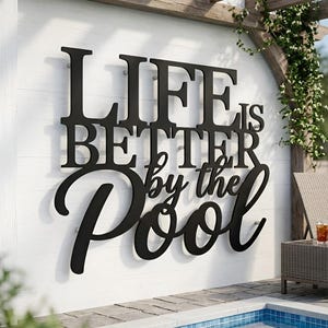 Könnte beinhalten: Schwarzes Metallschild mit der Aufschrift "LIFE IS BETTER by the POOL" an einer weißen Wand. Das Schild hat eine stilvolle Schriftart, ideal für die Außendekoration. Ein Pool und Gartenmöbel sind im Hintergrund zu sehen.