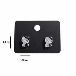 May include: A pair of Hello Kitty stud earrings. Each earring features a white and black cat design with a black bow. The earrings are displayed on a black card, with a height of 1.1 cm and a width of 0.08 cm.