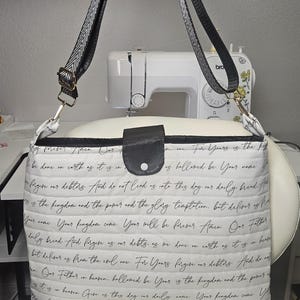 May include: White quilted fabric crossbody bag with black script text. The bag has a black leather flap closure and a black and white patterned strap. The bag is rectangular and appears to be handmade.