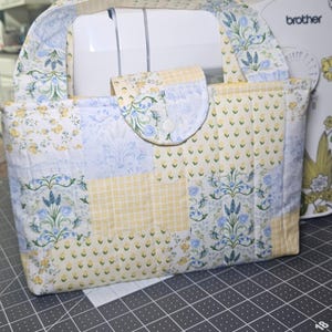 May include: A quilted sewing machine cover with a handle, featuring a patchwork design in yellow, blue, and white floral patterns. The cover has a flap closure with a button. A Brother sewing machine is visible in the background.