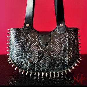 May include: Black and white snakeskin-patterned handbag with silver-tone spikes along the bottom and sides. The bag has a black leather handle and a flap closure. The background is a solid red.