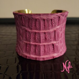 May include: A wide, magenta cuff bracelet with a textured, crocodile-like pattern. The interior of the bracelet is gold-toned. The bracelet is a bold fashion accessory.