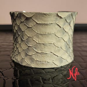 May include: A wide cuff bracelet with a textured, scaly pattern in shades of cream and grey. The bracelet has a curved shape and appears to be made of a reptile-like material. The design is reminiscent of snake or reptile skin.