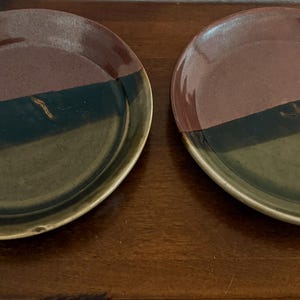 May include: Two handmade ceramic plates. Each plate is divided into three sections of color: burgundy, black, and olive green. The plates have a slightly irregular, organic shape and a glossy finish. The plates are displayed on a wooden surface.