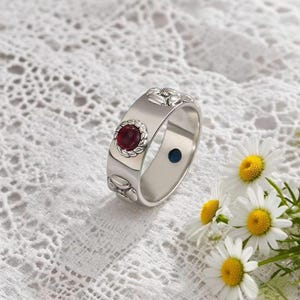 以下が含まれることがあります： 装飾的なベゼルにセットされた、大きくて丸い赤い宝石が付いたシルバーリング。リングは楕円形の模様の広いバンドと内側の小さな青い点があります。リングは白いレースの表面に、デイジーと一緒に展示されています。