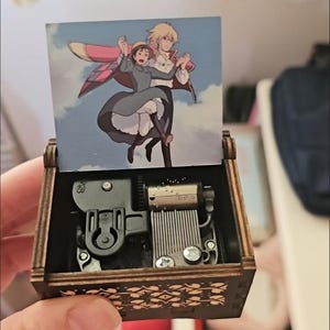 May include: A wooden music box featuring a colorful illustration of two animated characters against a blue sky. The box is dark brown with a decorative floral pattern on the sides. The internal mechanism is visible.