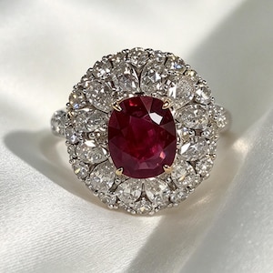 Puede incluir: Un anillo con un gran rubí rojo intenso de corte ovalado en el centro, asegurado por cuatro garras. El rubí está rodeado por un doble halo de numerosos diamantes blancos de corte pera y redondo engastados en una banda de metal plateado.