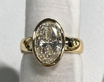May include: A gold ring featuring a large, oval-cut, clear faceted gemstone set in a smooth, polished bezel setting. The band is crafted from solid yellow gold with a high-shine finish, showcasing a clean, minimalist design.