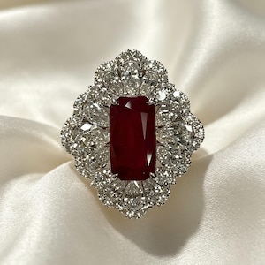 Peut inclure: Une bague ornée d'une grande pierre précieuse rectangulaire rouge foncé au centre, entourée d'un halo de multiples pierres blanches en forme de poire et rondes, serties dans une monture en métal argenté au bord extérieur festonné.