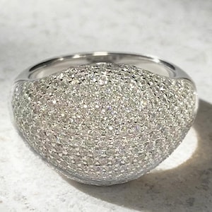 May include: A silver-toned dome ring featuring a dense pavé setting of numerous small, clear, sparkling stones covering the entire rounded surface. The band is smooth and polished, contrasting with the textured, reflective top of the ring.