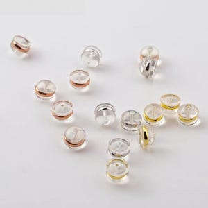May include: A collection of earring backs featuring a clear, circular silicone outer shell with a metallic center insert. The metal inserts are available in silver, gold, and rose gold tones, designed to secure earring posts firmly in place.