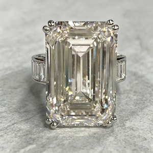 May include: A silver-toned ring featuring a large, rectangular emerald-cut center stone with a faceted surface. The band is accented with smaller, rectangular baguette-cut stones set along the sides, secured by four corner prongs.