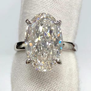 May include: A silver-toned ring featuring a large, oval-cut, clear faceted gemstone held in place by four prongs. The band is smooth and polished, showcasing a reflective metallic finish that complements the brilliant, multi-faceted stone.