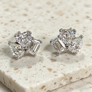 Könnte beinhalten: Ein Paar Diamant-Ohrstecker. Jeder Ohrring hat drei klare, funkelnde Diamanten in verschiedenen Formen: rund, birnenförmig und Smaragd-Schliff. Die Ohrringe sind in einem silberfarbenen Metall gefasst.