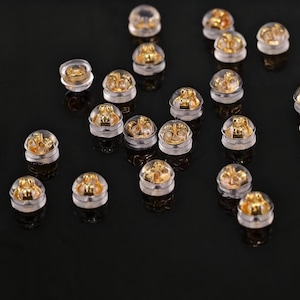 Puede incluir: Un conjunto de cierres para pendientes, cada uno con un enganche de metal dorado encapsulado en una almohadilla transparente de silicona o plástico en forma de cúpula, diseñados para asegurar los pendientes en su lugar.