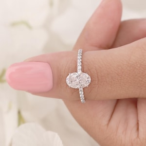 May include: A diamond engagement ring with an oval-cut center stone. The band is encrusted with small, sparkling diamonds. The ring is held up against a soft pink background.