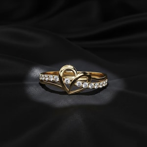 May include: A gold ring with a heart-shaped design. The ring features a band with small, clear stones and a heart-shaped centerpiece. The ring is set against a black background, highlighting its details and shine.