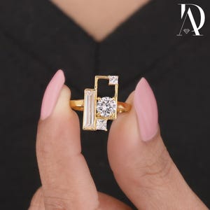 Puede incluir: Un anillo de oro con un diseño geométrico, con una gema clara grande y redonda en el centro, rodeada de gemas claras más pequeñas, rectangulares y cuadradas. El anillo se sostiene entre dos dedos sobre un fondo negro.