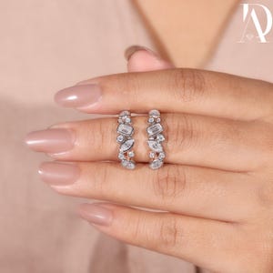 May include: Silver hoop earrings adorned with clear gemstones. The earrings showcase a mix of rectangular and marquise-cut stones, creating a delicate design. The earrings are held up by a hand with painted nails. The earrings are a stylish accessory.