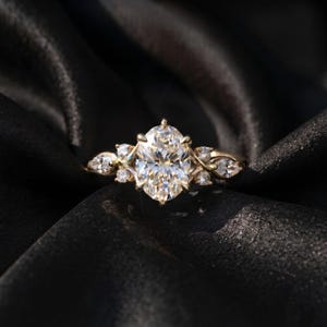 May include: A gold ring featuring a large oval-cut center stone surrounded by smaller round and marquise-cut stones. The band has a delicate, intricate design with multiple prongs securing the central gemstone in a classic, elegant arrangement.
