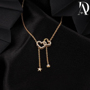 May include: A gold necklace featuring two heart-shaped pendants adorned with small, clear gemstones. The necklace has a fine chain and includes a dangling star and a small round gem. The backdrop is a dark, textured fabric.