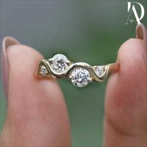 Könnte beinhalten: Ein goldener Ring mit einem einzigartigen Wellendesign, besetzt mit drei runden, brillantgeschliffenen Diamanten. Der Ring wird zwischen Fingern gehalten, was die komplizierten Details der Fassung und das Funkeln der Edelsteine hervorhebt. Der Ring ist ein Schmuckstück.
