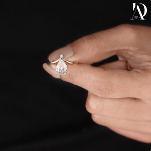 May include: A gold ring featuring a pear-shaped, clear gemstone. The ring has a delicate, curved band and a small, pointed accent above the main stone. The ring is held against a dark background.