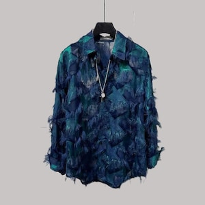 Puede incluir: Una camisa de manga larga con cuello, en tonos turquesa y azul marino, adornada con un diseño texturizado con flecos. Se ve un collar plateado con un colgante. La camisa se exhibe en una percha negra sobre un fondo neutro.