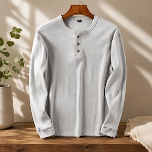 Könnte beinhalten: Ein hellgraues, langärmeliges Henley-Shirt mit vertikaler Rippenstruktur, rundem Halsausschnitt und einer Knopfleiste mit drei dunkelbraunen Knöpfen. Das Kleidungsstück hängt an einem Holzkleiderbügel und zeigt seine schlichte Textur.