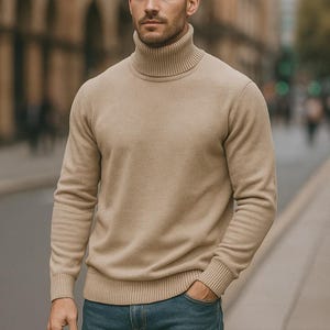 May include: A man wearing a tan turtleneck sweater and blue jeans stands outdoors. The sweater has a ribbed collar, cuffs, and hem. The jeans are a classic blue denim. The background is a blurred city street.