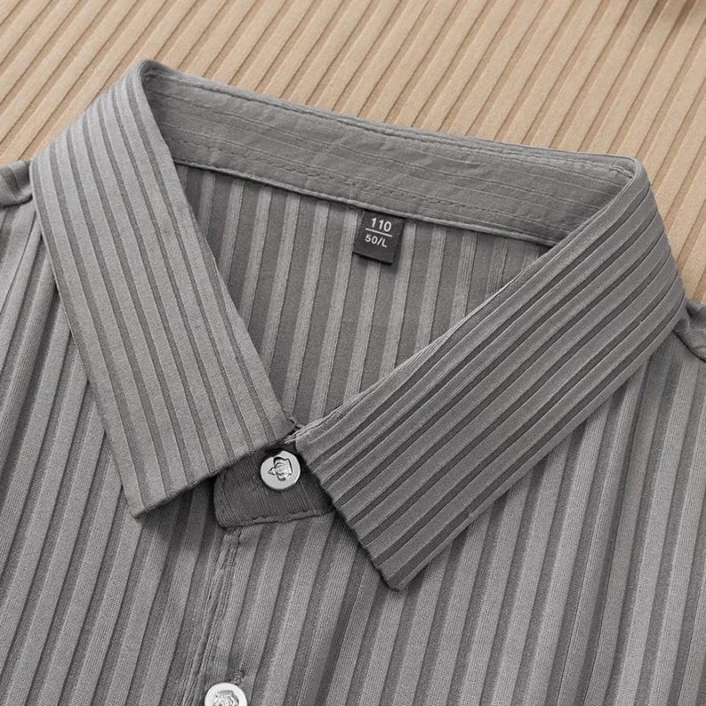May include: Close-up of a gray collared shirt with vertical ribbed detailing. The shirt has a button-down closure and a small tag with the numbers "110" and "50/L". The fabric has a subtle sheen.