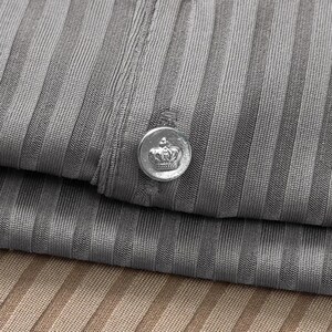 May include: Close-up of a gray ribbed fabric with a silver button featuring a crown design. The button is attached to the folded fabric, with a second layer of fabric visible below in a tan color.