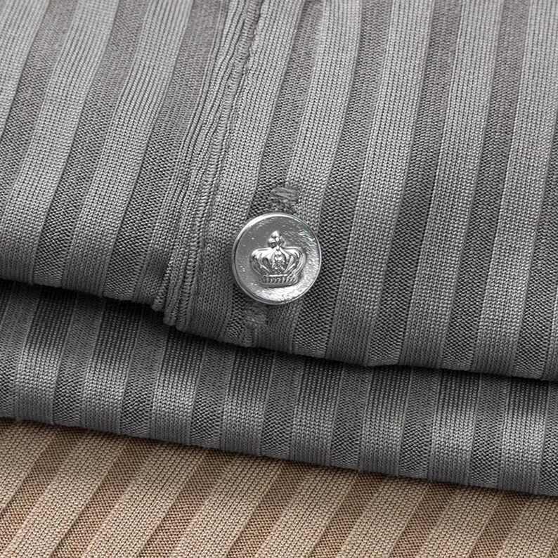May include: Close-up of a gray ribbed fabric with a silver button featuring a crown design. The button is attached to the folded fabric, with a second layer of fabric visible below in a tan color.