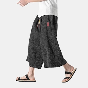 May include: Dark gray wide-leg pants with a textured appearance. The pants have a small red emblem on the pocket and a drawstring. The person is wearing black sandals with a light brown sole.