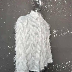 Puede incluir: Una camisa blanca de manga larga con cuello y un dise&ntilde;o texturizado &uacute;nico. La camisa est&aacute; cubierta de un material plumoso y con flecos, creando un efecto visual llamativo. La prenda se exhibe en un maniqu&iacute;.