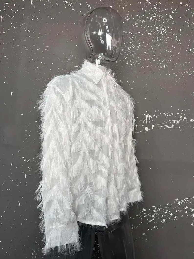 Puede incluir: Una camisa blanca de manga larga con cuello y un dise&ntilde;o texturizado &uacute;nico. La camisa est&aacute; cubierta de un material plumoso y con flecos, creando un efecto visual llamativo. La prenda se exhibe en un maniqu&iacute;.