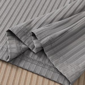 May include: Close-up of a gray ribbed long-sleeve top. The fabric has a textured, vertical striped pattern. The sleeves are folded over, revealing the stitching and the inside of the garment. The background is a light brown ribbed fabric.
