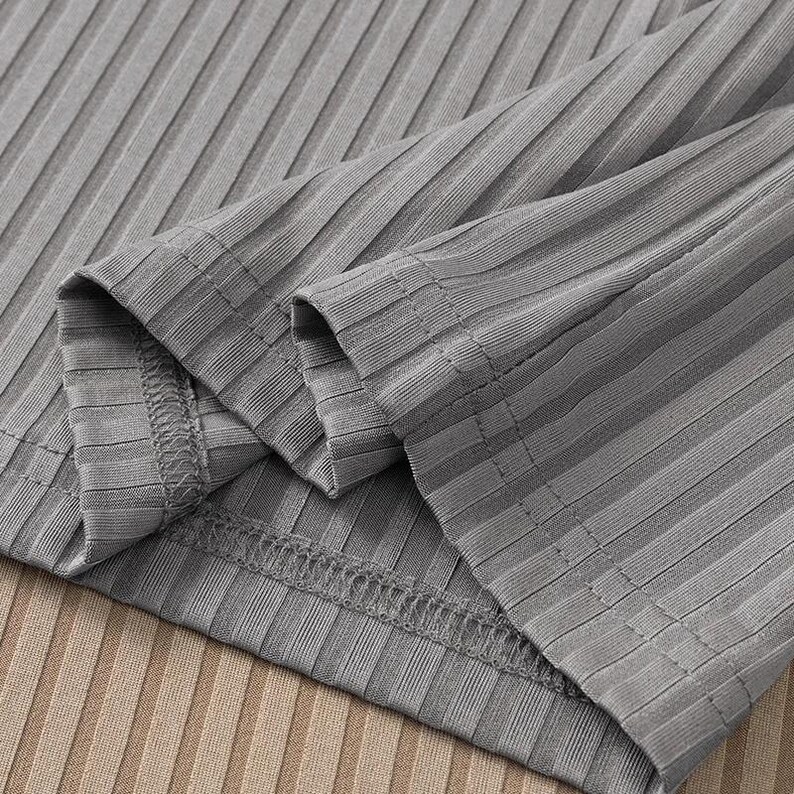May include: Close-up of a gray ribbed long-sleeve top. The fabric has a textured, vertical striped pattern. The sleeves are folded over, revealing the stitching and the inside of the garment. The background is a light brown ribbed fabric.