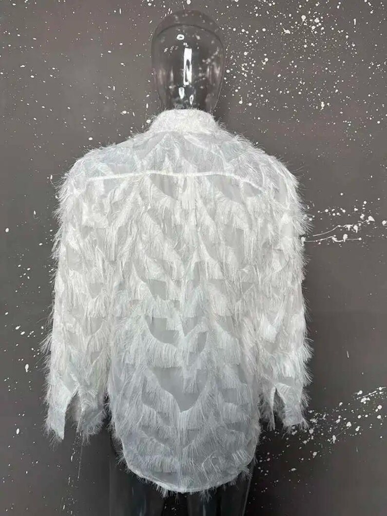 Puede incluir: Camisa blanca de manga larga con un dise&ntilde;o texturizado y plumoso. La camisa tiene un cuello cl&aacute;sico y una botonadura frontal. La tela parece ligera y semitransparente, con una textura &uacute;nica con flecos.