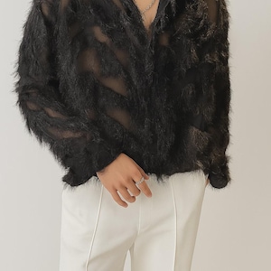 Puede incluir: Camisa negra transparente de manga larga con textura difusa. La camisa tiene cuello y se combina con pantalones blancos. Se ve un collar de cadena plateada. El estilo general es moderno y &uacute;nico.