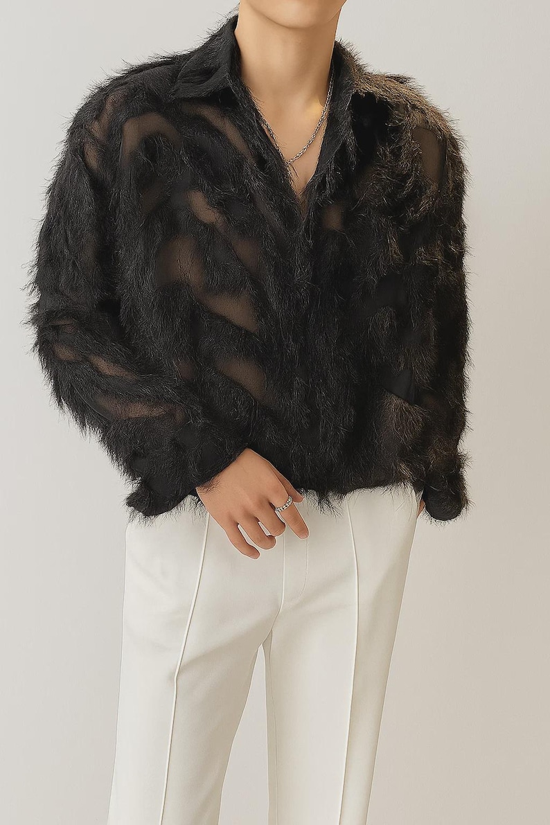 Puede incluir: Camisa negra transparente de manga larga con textura difusa. La camisa tiene cuello y se combina con pantalones blancos. Se ve un collar de cadena plateada. El estilo general es moderno y &uacute;nico.