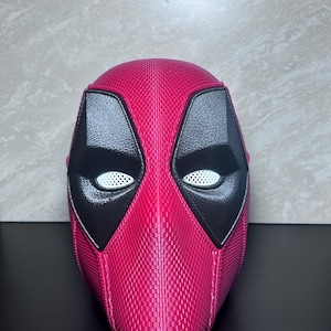 May include: A pink and black Deadpool mask. The mask features a textured pink surface with black eye and mouth details. The eyes have white mesh inserts. The mask is a full-face design.