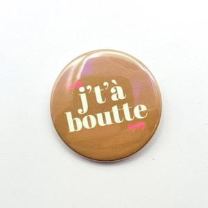 May include: A circular, bronze-toned badge featuring the words "j't'à boutte" in white. Pink details are visible. The badge is set against a white backdrop.