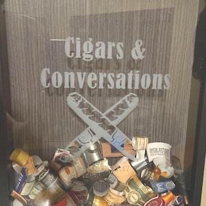 May include: A framed shadow box with a wooden background and the text "Cigars & Conversations". The box contains a collection of cigar bands and a decorative design of crossed cigar cutters.