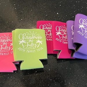 May include: A selection of drinks can coolers in shades of pink, green, and purple. Each cooler displays the text "Christmas in July" with palm tree designs and "ROTH FAMILY 2024" in white, ideal for a summer event.