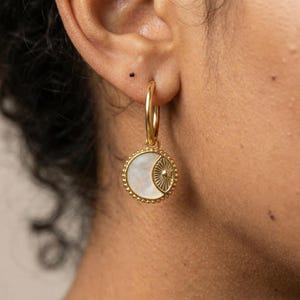 May include: Gold hoop earrings with a circular pendant. The pendant has a white, iridescent mother-of-pearl background and a gold sunburst design. The pendant is framed with a beaded gold border. These earrings are a jewellery accessory.