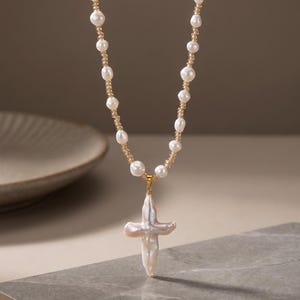 Può includere: Una collana composta da una fila di perle d'acqua dolce bianche alternate a piccole perline dorate traslucide. La collana presenta un grande ciondolo in perla barocca dalla forma irregolare a croce su una montatura dorata.